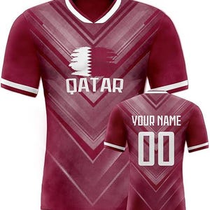 May include: Maroon soccer jersey with white accents. The front features the word "QATAR" below a graphic. The back has the text "YOUR NAME" above the number "00". The jersey has a V-neck collar and white stripes on the sleeves.