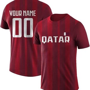 May include: Two maroon t-shirts with vertical stripes. One shirt displays "YOUR NAME" and the number "00" in white. The other shirt has "QATAR" in white, with a small flag graphic. Both shirts have a crew neck.