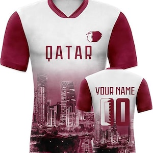 May include: White and maroon soccer jersey with the word "QATAR" in maroon block letters. The jersey features a cityscape design and the text "YOUR NAME" and the number "00" on the back.