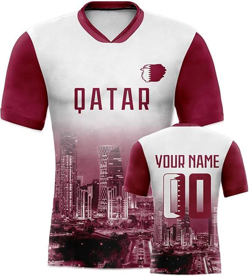 May include: White and maroon soccer jersey with the word "QATAR" in maroon block letters. The jersey features a cityscape design and the text "YOUR NAME" and the number "00" on the back.