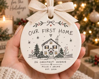 May include: A round white ceramic ornament with a beige fabric ribbon bow. The front features a black ink illustration of a house with a chimney, trees, and a fence. Text reads: OUR FIRST HOME, 26 CHESTNUT AVENUE, MILLIE + ARCHIE, EST. 2026.