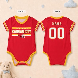 Puede incluir: Un body para beb&eacute; de color rojo con ribetes amarillos, cuello en V y mangas cortas. El frente muestra el texto KANSAS CITY FOOTBALL con un gr&aacute;fico de casco. La parte posterior presenta la palabra NAME sobre el n&uacute;mero 00 en letras blancas.