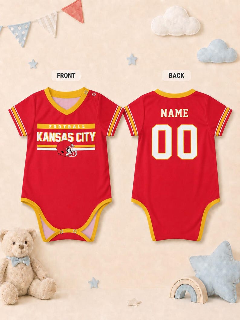 Puede incluir: Un body para beb&eacute; de color rojo con ribetes amarillos, cuello en V y mangas cortas. El frente muestra el texto KANSAS CITY FOOTBALL con un gr&aacute;fico de casco. La parte posterior presenta la palabra NAME sobre el n&uacute;mero 00 en letras blancas.