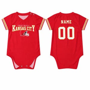 May include: A red short-sleeved baby bodysuit featuring the text KANSAS CITY FOOTBALL and a helmet graphic on the front. The back displays the word NAME and the number 00 in white lettering. The sleeves have yellow and white stripes.