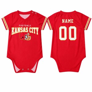 May include: A red short-sleeved baby bodysuit featuring the text KANSAS CITY FOOTBALL and a helmet graphic on the front. The back displays the word NAME above the number 00 in white with yellow outlines. The sleeves have yellow and white stripes.