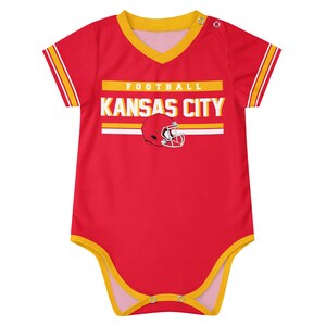 Puede incluir: Un body de beb&eacute; rojo de manga corta con ribetes amarillos en el cuello, las mangas y las aberturas de las piernas. El frente presenta el texto FOOTBALL KANSAS CITY en blanco y amarillo, junto a un gr&aacute;fico de un casco de f&uacute;tbol rojo.