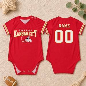 May include: A red baby bodysuit featuring a Kansas City football graphic on the front and the text NAME with the number 00 on the back. The short-sleeve garment has a V-neckline, white and yellow sleeve stripes, and snap closures at the shoulder and crotch.