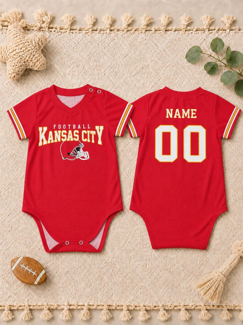 May include: A red baby bodysuit featuring a Kansas City football graphic on the front and the text NAME with the number 00 on the back. The short-sleeve garment has a V-neckline, white and yellow sleeve stripes, and snap closures at the shoulder and crotch.