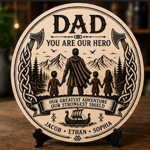 Może przedstawiać: Okrągła drewniana tablica z napisem "DAD" na górze i tekstem "YOU ARE OUR HERO". Tablica przedstawia motyw wikingów z ilustracją rodziny, toporami i słowami "OUR GREATEST ADVENTURE OUR STRONGEST SHIELD".