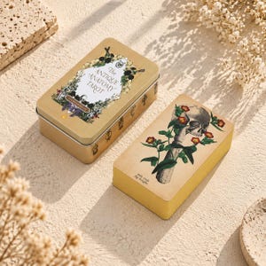 May include: A rectangular gold-coloured metal tin labelled The Antique Anatomy Tarot with floral illustrations and a skull, alongside a deck of tarot cards featuring a skull entwined with green vines and red flowers, marked with the text suit rods fig. knight.