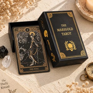 Puede incluir: Una caja de cartas de tarot negra con el texto dorado The Marigold Tarot y un emblema de calavera, junto a una carta de tarot con la ilustración de un esqueleto etiquetada como The Fool, decoradas con líneas doradas y motivos botánicos.
