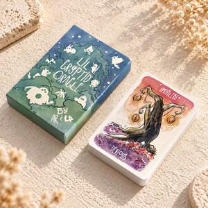May include: A rectangular deck box titled Lil Cryptid Oracle by Ar & La in blue and green with white ghost illustrations, alongside a single rectangular oracle card featuring a dark tree with glowing lanterns and the text Devious and Umdaledi.