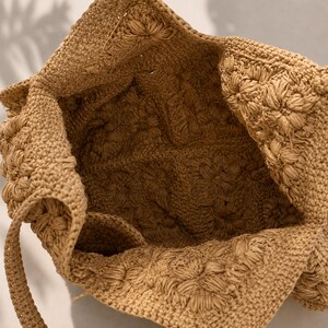 May include: A tan crochet tote bag featuring a square-based construction with a textured floral pattern throughout. The bag has a wide open top and a single integrated strap, showcasing a consistent light brown yarn weave and detailed stitching.