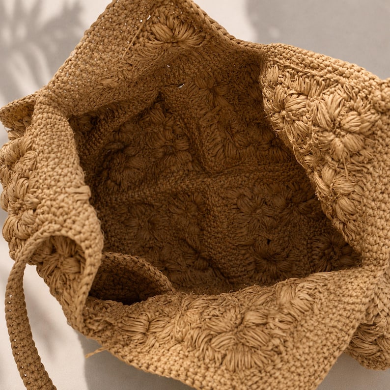May include: A tan crochet tote bag featuring a square-based construction with a textured floral pattern throughout. The bag has a wide open top and a single integrated strap, showcasing a consistent light brown yarn weave and detailed stitching.