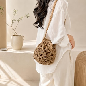 May include: A tan macrame bucket bag featuring an intricate knotted pattern, a braided shoulder strap, and a drawstring closure secured with two dark brown wooden beads. The bag is crafted from natural cord and has a rounded, soft-sided structure.