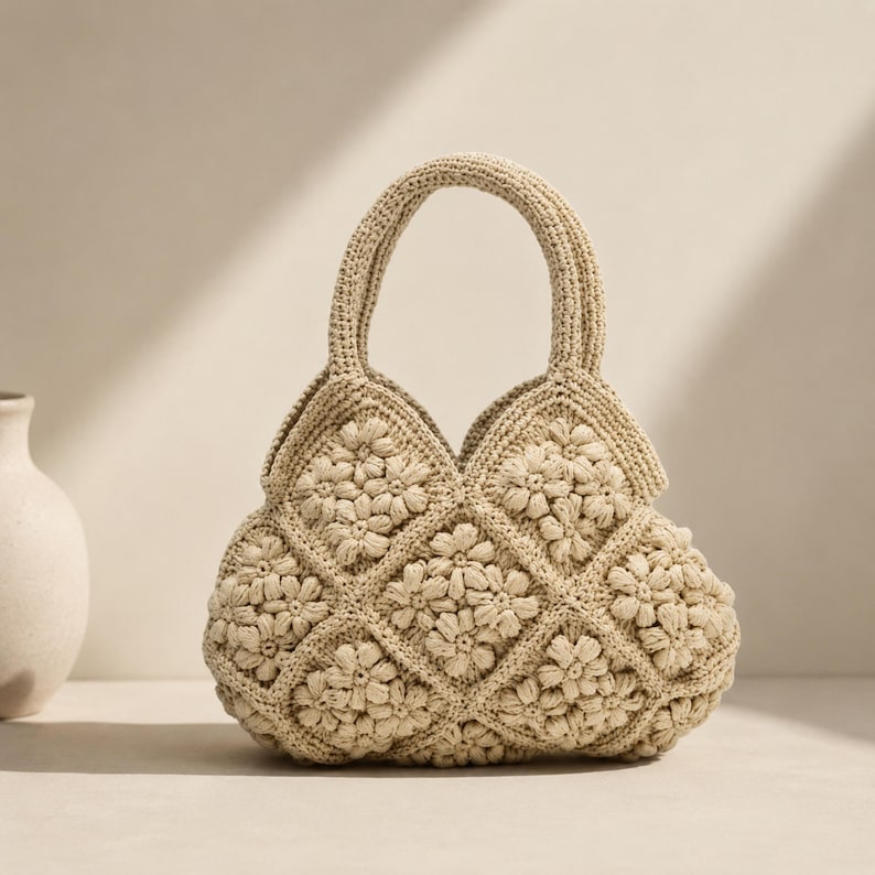 May include: A beige crochet handbag featuring a repeating diamond pattern composed of textured floral motifs. The bag has a structured top handle and a scalloped upper edge, crafted from a light-coloured yarn with a consistent knit texture throughout.