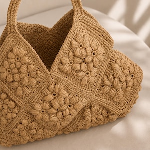 May include: A tan crochet handbag constructed from multiple square panels featuring a raised floral motif. The bag has a wide, open top and a single integrated handle, showcasing a textured, hand-stitched appearance in a uniform light brown colour.