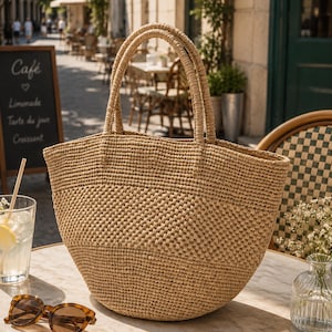 Può includere: Una grande borsa tote in paglia intrecciata color beige con due manici robusti. La borsa presenta un motivo a trama di cesto su tutto il corpo, creando una forma strutturata ma flessibile. Il materiale è di un colore marrone chiaro naturale.