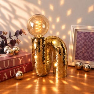 May include: A gold disco ball table lamp with a curved, tubular base covered in small reflective square mirrors. The lamp features a large, round, amber-tinted Edison-style light bulb with a visible glowing filament, casting light patterns.