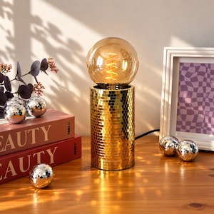 Peut inclure: Une lampe de table cylindrique recouverte de petites tuiles miroir dorées, surmontée d'une grande ampoule en verre teinté ambre avec un filament en spirale visible. La base de la lampe reflète la lumière, créant un effet scintillant.
