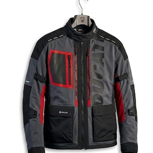 May include: A grey and black motorcycle jacket with red detailing and the word "DUCATI" printed on the front. The jacket features multiple pockets, zips, and the "GORE-TEX" logo. It is suspended from a hanger.