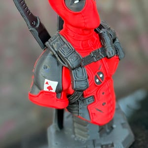 May include: A red and black Deadpool bust figurine featuring a character wearing a tactical harness, a shoulder pad with an ace of spades playing card emblem, and two swords strapped to the back, all mounted on a grey hexagonal base.