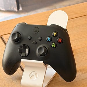 Pode incluir: Um controle Xbox preto em um suporte branco. O controle tem botões coloridos (verde, vermelho, azul, amarelo) e joystiques pretos. O suporte tem o logotipo da Xbox.