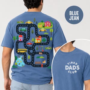 May include: Blue jean-colored t-shirt with a whimsical road map design on the back, featuring colorful cars, houses, trees, and a school bus. The front of the shirt displays the text "TIRED DADS CLUB" in white.