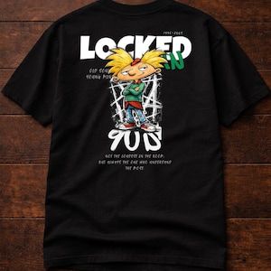 Peut inclure: T-shirt noir avec un personnage de dessin animé aux cheveux jaunes et une veste verte. Le t-shirt affiche le mot "LOCKED" au-dessus du personnage, avec le texte "OLD SOUL YOUNG MIND" et "90s" en dessous. Le t-shirt a également le texte "NOT THE LOUDEST IN THE ROOM, BUT ALWAYS THE ONE WHO UNDERSTAND THE MOST."