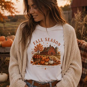 Peut inclure: Un t-shirt blanc avec une illustration représentant une grange rouge, un panier de citrouilles et des feuilles d'automne. Le texte indique FALL SEASON et GOOD THINGS TAKE TIME EST 2024. Porté sous un gilet en maille beige, jean bleu et ceinture marron.