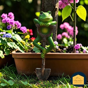 May include: A green frog garden ornament holding a shovel, placed in a flower pot. The frog has a smiling face and is leaning on the shovel. The garden ornament is surrounded by colourful flowers and greenery.