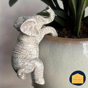 Puede incluir: Figura de elefante blanco envejecido trepando por el lateral de una maceta de cerámica. El elefante tiene una textura detallada y está posicionado como si estuviera alcanzando la planta del interior. La maceta es clara con un borde marrón.