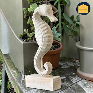 May include: A carved wooden seahorse sculpture. The seahorse is light-coloured and stands upright on a rectangular wooden base. The sculpture has detailed carvings, showcasing the seahorse's form. The background includes potted plants.