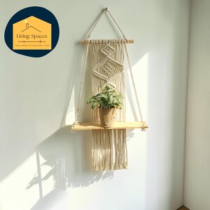 Puede incluir: Estante para plantas de macramé con un estante de madera y una planta en maceta. El estante está suspendido por una cuerda y presenta un diseño geométrico. El estante está hecho de cuerda y madera de color natural, creando una estética bohemia.