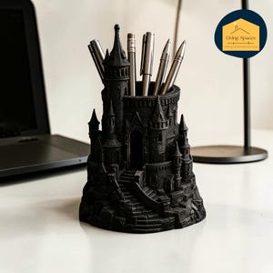 May include: Black castle-shaped pen holder with multiple turrets, towers, and a central cylindrical compartment holding pens and pencils. The pen holder is made of a dark material and sits on a white surface. The Living Spaces logo is in the background.