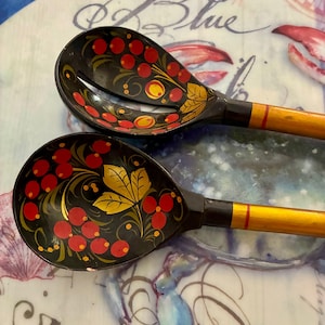 May include: Two wooden spoons with black bowls featuring hand-painted red berries and gold leaves. One spoon is a solid bowl, while the other is a slotted serving spoon. Both have long, smooth, yellow-toned handles with a thin red accent stripe.