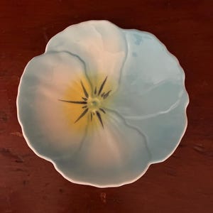 Puede incluir: Un plato de cerámica con forma de flor. El plato tiene un exterior azul claro con un centro amarillo y azul oscuro. El plato tiene un borde festoneado.