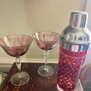 Può includere: Uno shaker per cocktail rosso con la parte superiore argentata e due bicchieri da coupé rosa abbinati. I bicchieri e lo shaker presentano disegni floreali incisi. Lo shaker ha un motivo a rombi. Il set è esposto su una superficie di legno scuro.