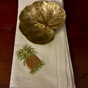 以下が含まれることがあります： 金色の葉の形をしたお皿が、刺繍されたパイナップルのデザインの白いリネンナプキンに置かれています。ナプキンには装飾的なステッチの縁があり、トロピカルなテーマを作り出しています。