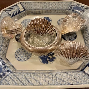 Könnte beinhalten: Eine Sammlung dekorativer Artikel, darunter eine Glasschale mit silbernem Rand und muschelförmigem Aufsatz, muschelförmige Untersetzer und ein Stapel kleiner Untersetzer, alles präsentiert auf einem blau-weiß gemusterten Keramiktablett.