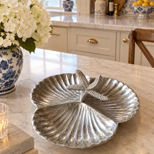 May include: A silver-toned metal serving dish shaped like a three-sectioned scallop shell, featuring a textured starfish accent in the center. The dish has a metallic finish with visible ridges radiating from the center to the scalloped edges.