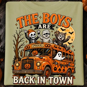 Puede incluir: Una camiseta verde oliva claro con un gr&aacute;fico de un autob&uacute;s escolar naranja con un esqueleto, una momia y un hombre lobo en el techo. El texto dice THE BOYS ARE BACK IN TOWN y EPOEKRS. Un fantasma est&aacute; en la ventana y calabazas decoran el autob&uacute;s.