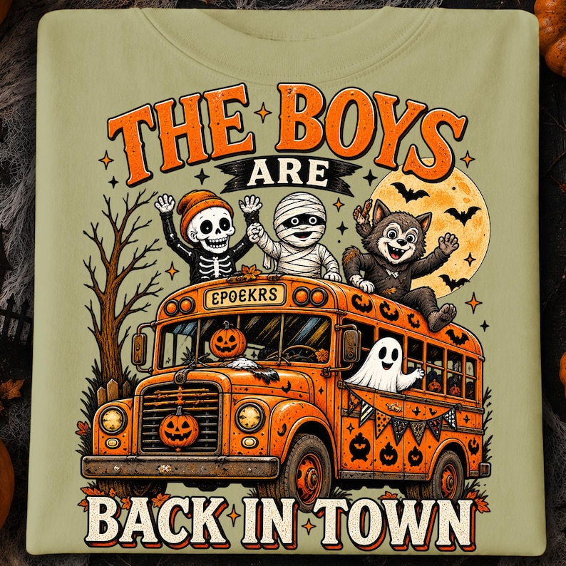 Puede incluir: Una camiseta verde oliva claro con un gr&aacute;fico de un autob&uacute;s escolar naranja con un esqueleto, una momia y un hombre lobo en el techo. El texto dice THE BOYS ARE BACK IN TOWN y EPOEKRS. Un fantasma est&aacute; en la ventana y calabazas decoran el autob&uacute;s.