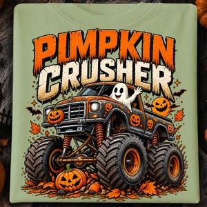 Puede incluir: Una camiseta verde claro con un gr&aacute;fico de un cami&oacute;n monstruo de neum&aacute;ticos grandes, decorado con motivos de calabazas y calaveras naranjas. Un fantasma blanco est&aacute; sentado en el asiento del conductor. El texto PUMPKIN CRUSHER aparece en letras grandes de color naranja y beige desgastadas sobre el cami&oacute;n.
