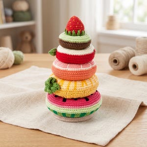 Könnte beinhalten: Ein farbenfrohes Häkel-Steckspielzeug in Form eines Fruchtturms. Das Spielzeug hat eine Wassermelonenbasis, eine gelbe Ananasschicht, eine orangefarbene Schicht, eine rote Erdbeerschicht und eine Erdbeerspitze. Das Spielzeug ist handgefertigt und liegt auf einem neutralen Leinentuch.