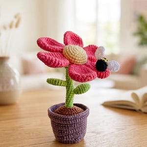 May include: A crocheted flower in a purple pot. The flower features bright pink petals, a yellow centre, and a green stem with leaves. A small bee with black and white wings is attached. The pot is filled with brown yarn.