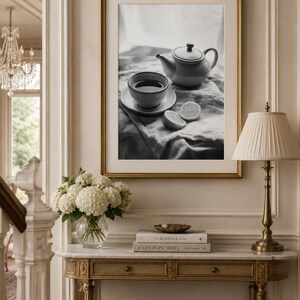 Puede incluir: Una impresión de arte mural enmarcada en blanco y negro que muestra una tetera, una taza de té sobre un platillo y dos mitades de limón sobre una tela drapeada. La obra está en un marco dorado con un paspartú blanco.