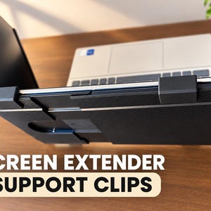 May include: A black, textured screen extender support clip attached to the side of a laptop. The device features a rigid, rectangular frame with a carbon fibre-like pattern and two prominent, square-shaped mounting brackets securing the display.