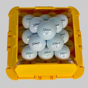 May include: A yellow plastic golf ball storage container filled with white Titleist golf balls. The golf balls are arranged in neat rows and have the number 1, 2, or 4 printed on them.