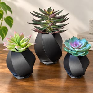 May include: Three matte black geometric planters with a faceted, twisted design. Each planter holds a different artificial succulent: one with light green and pink leaves, one with dark green and purple leaves, and one with blue-green leaves.